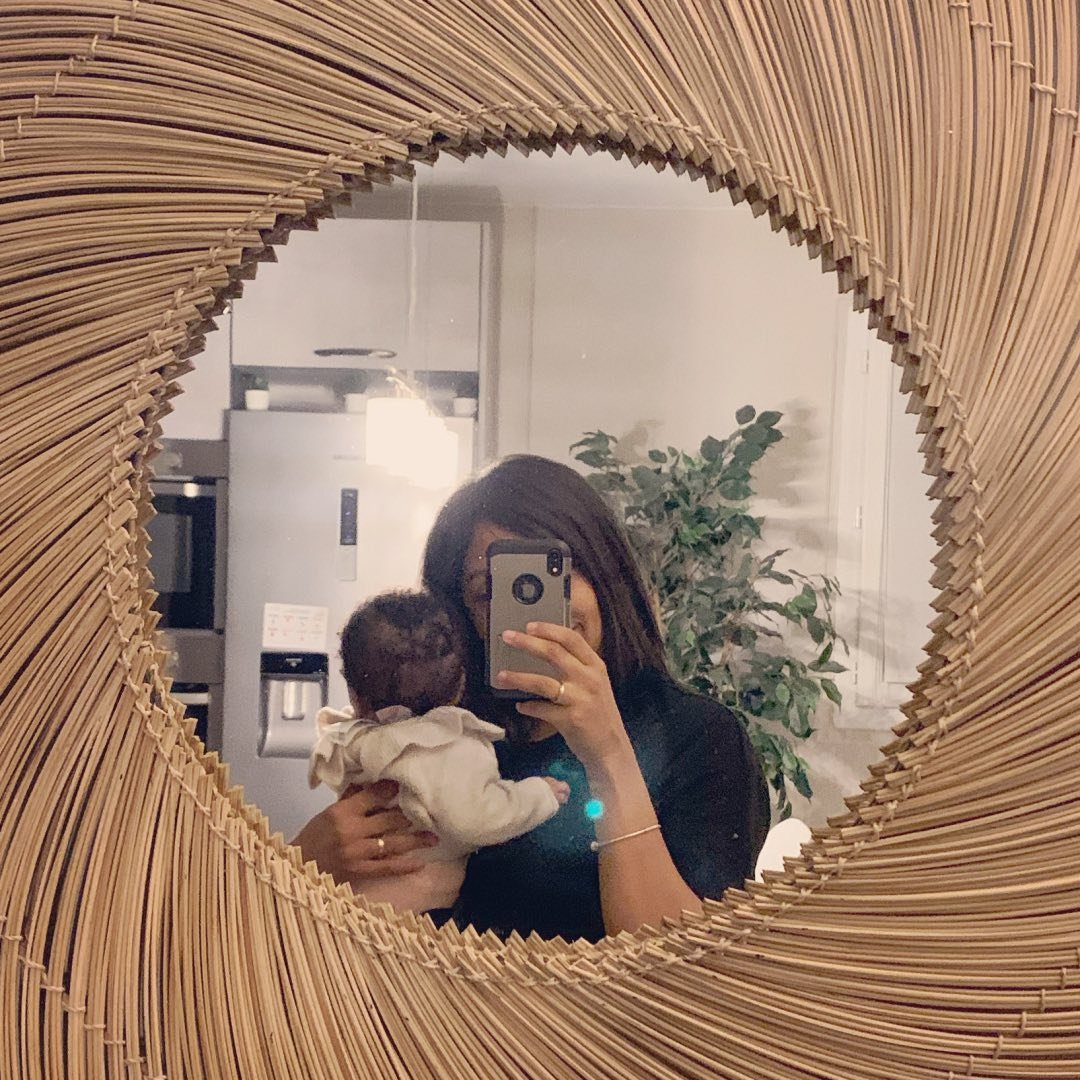 Woman holding a baby in a circular woven wall mirror.
